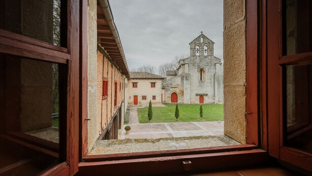 Vista attraverso una finestra aperta su un cortile con un edificio, una torre campanaria, una zona pavimentata e qualche albero.