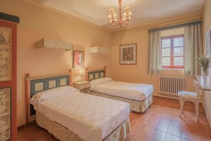 Una habitación con dos camas, una ventana con cortinas, un radiador, un escritorio con silla y una araña.