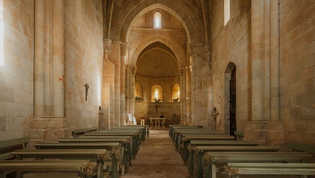 Un interior con techos abovedados y paredes de piedra, hileras de bancos y un altar central.