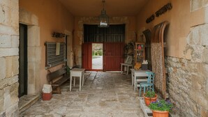 Un pasillo con suelo de piedra, un banco, una mesa y sillas, que conduce a una puerta de madera.