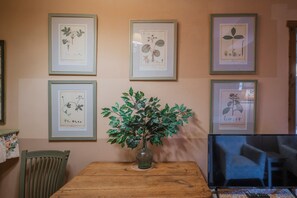 Une pièce avec une table en bois, une chaise verte, un vase contenant des feuilles vertes et, au mur, des illustrations botaniques encadrées.