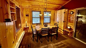 A wooden cabin interior with a dining table, chairs, a chandelier, and a window with blinds. - The Nordico Point Cabin (Ely)