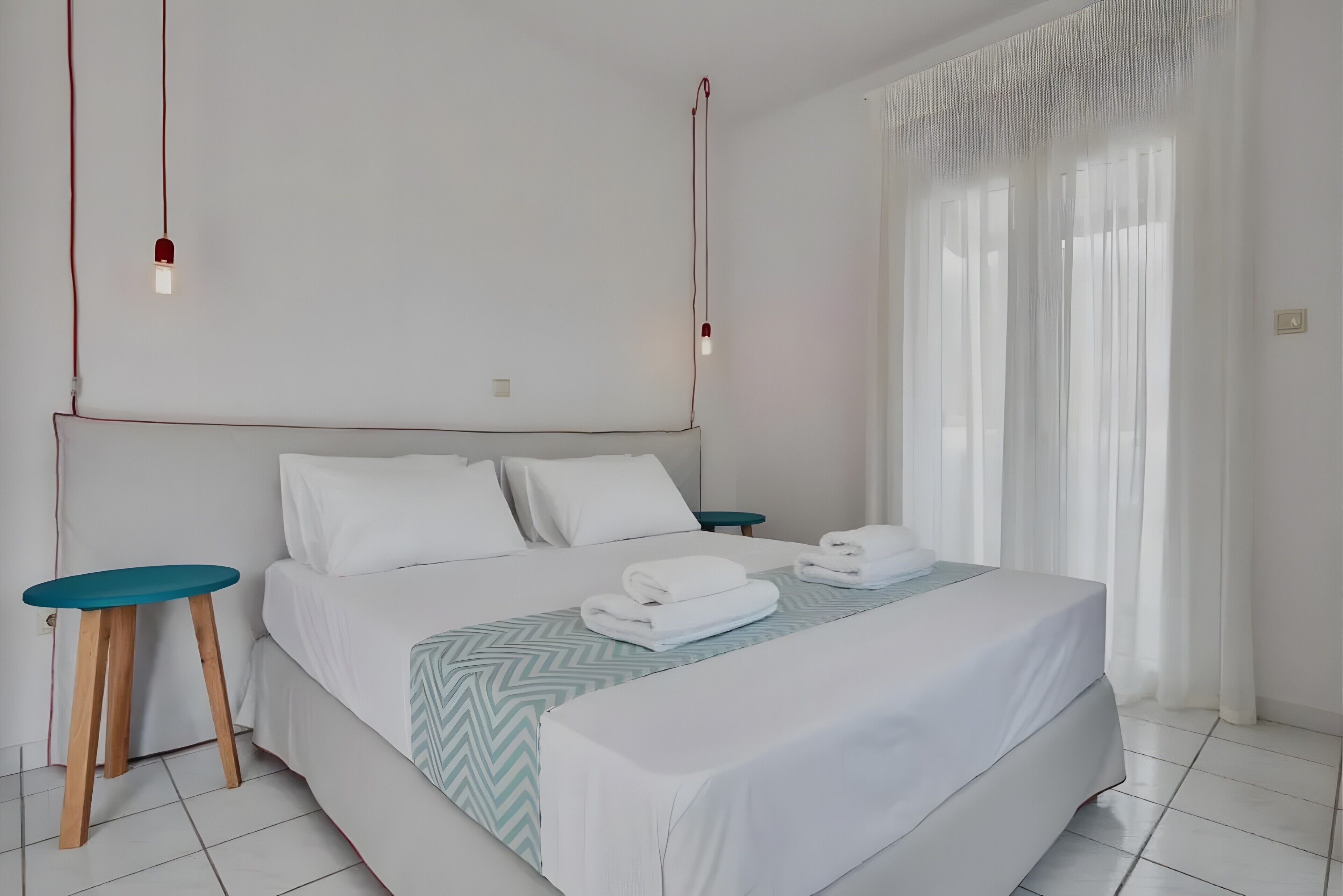 A neatly made bed with white linens, a green patterned bedspread, and two folded towels. There are two bedside tables with wooden legs and a small round table. The room has white walls, a large window with sheer curtains, and a ceiling light fixture.