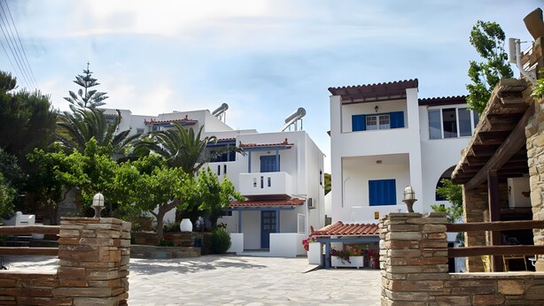 A courtyard with stone flooring, white buildings with blue shutters, and a stone wall with a gate.