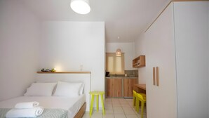 A hotel room with a bed, a small table, a chair, a wardrobe, and a kitchenette in the background.