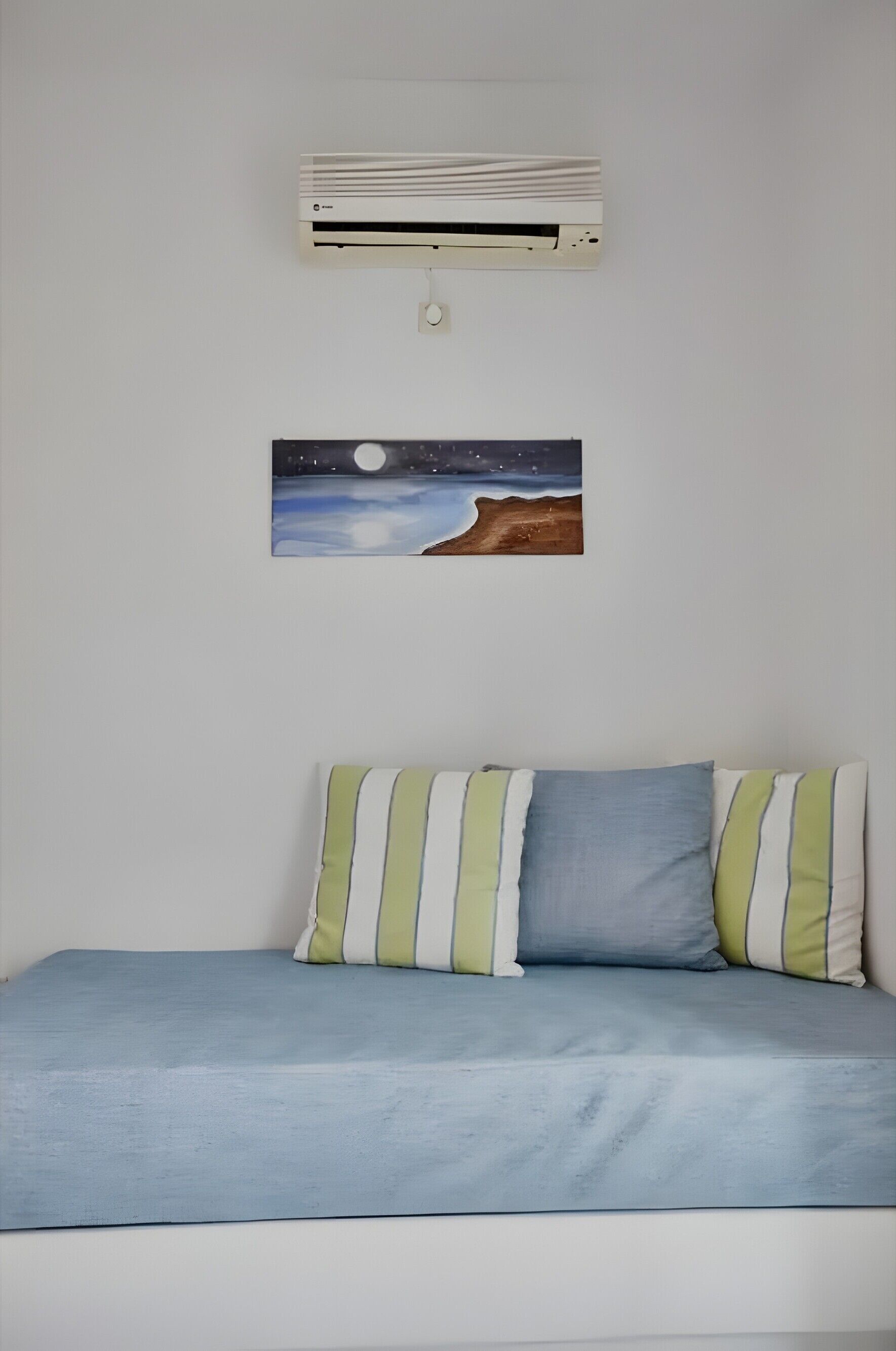 A bed with striped pillows, a wall-mounted air conditioner, and a painting of a night scene.