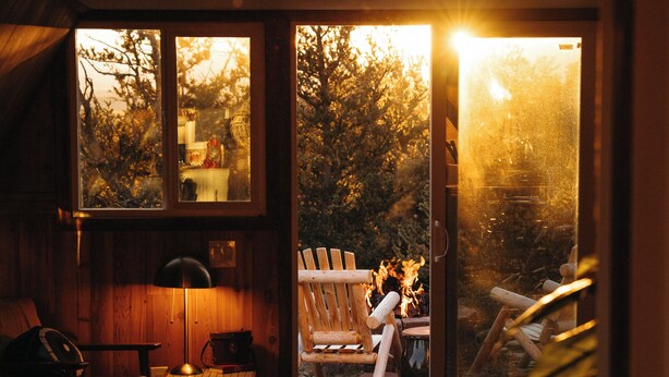 Una habitación acogedora con detalles de madera, un escritorio con ordenador y una silla. Hay un ventanal con vistas a árboles y una fogata en el exterior.
