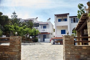 A stone-paved courtyard with white buildings, a stone wall, and a wooden pergola with flowers.