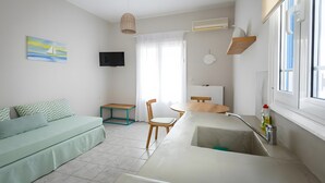 A compact living space with a sofa, a dining table, a kitchen area with a sink, a television, and a window with curtains. - Studio Apartment 'Semi Basement -Superior Apartment' with Mountain View and Air Conditioning (Andros)