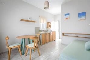 A modern kitchen with wooden cabinets, a dining area with a round table and chairs, a bed with a headboard, and a view of the sea.