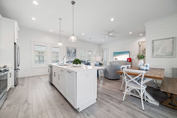A modern kitchen with white cabinetry and a central island, a dining area with a wooden table and chairs, and a large window with blinds.