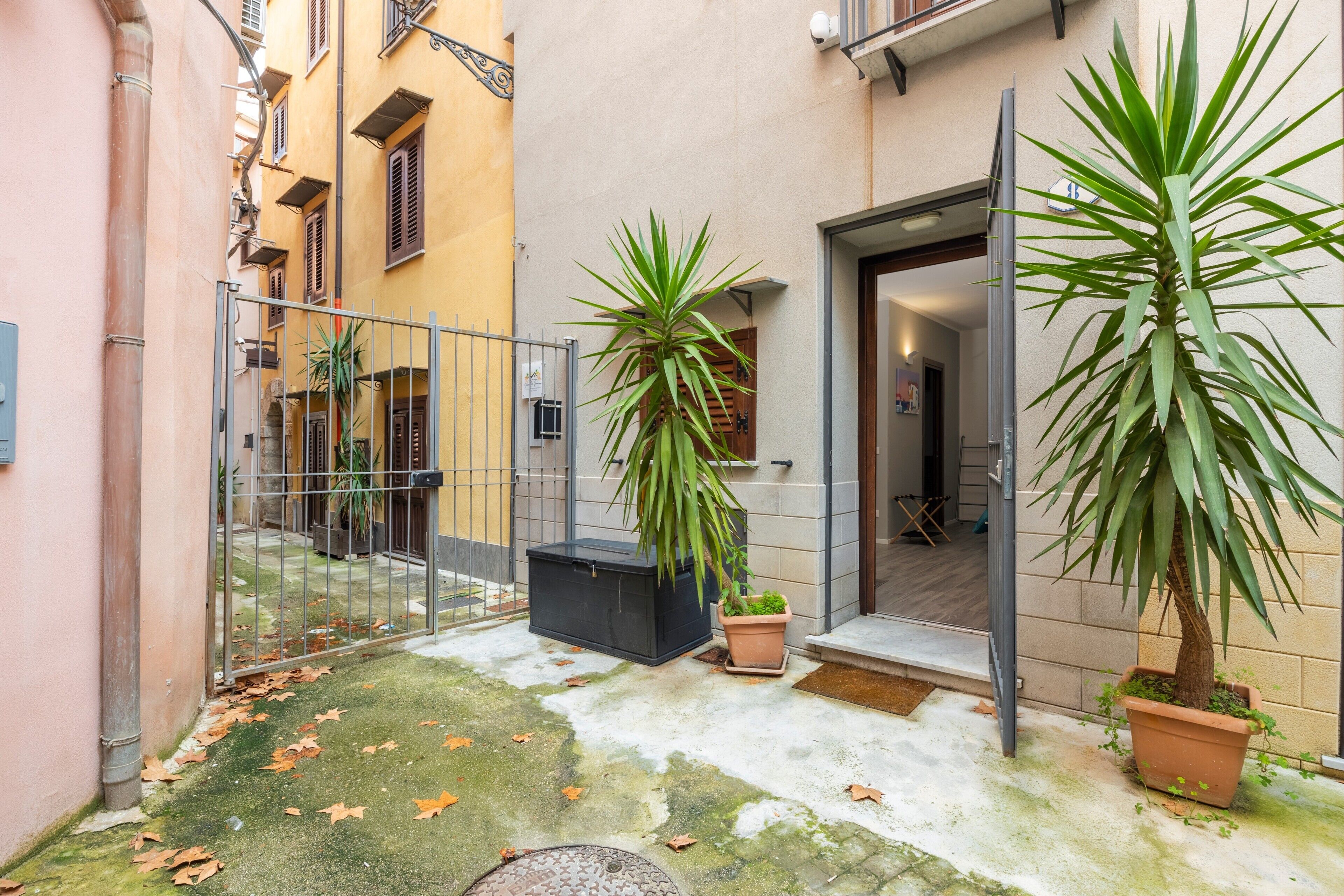 Un cortile con un cancello in metallo, piante in vaso, un pavimento in cemento e l'ingresso di un edificio.