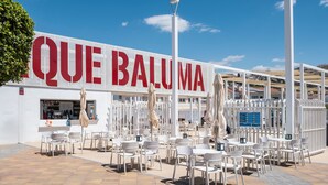 A cafe with outdoor seating, a large sign reading 'QUE BALUMA,' and a clear blue sky. - Apartment 'Casa Santo Front Beach' with Private Terrace and Wi-Fi (Cala del Moral)
