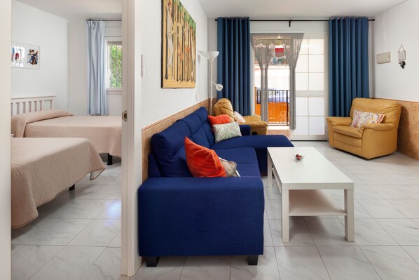 Ein Zimmer mit einem blauen Sofa, einem gelben Sessel, einem weißen Couchtisch und einem Bett mit einer beigefarbenen Decke.