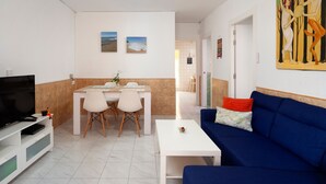A living room with a blue sofa, a white coffee table, a TV, and a dining area with a table and chairs. - Apartment 'Casa Santo Front Beach' with Private Terrace and Wi-Fi (Cala del Moral)