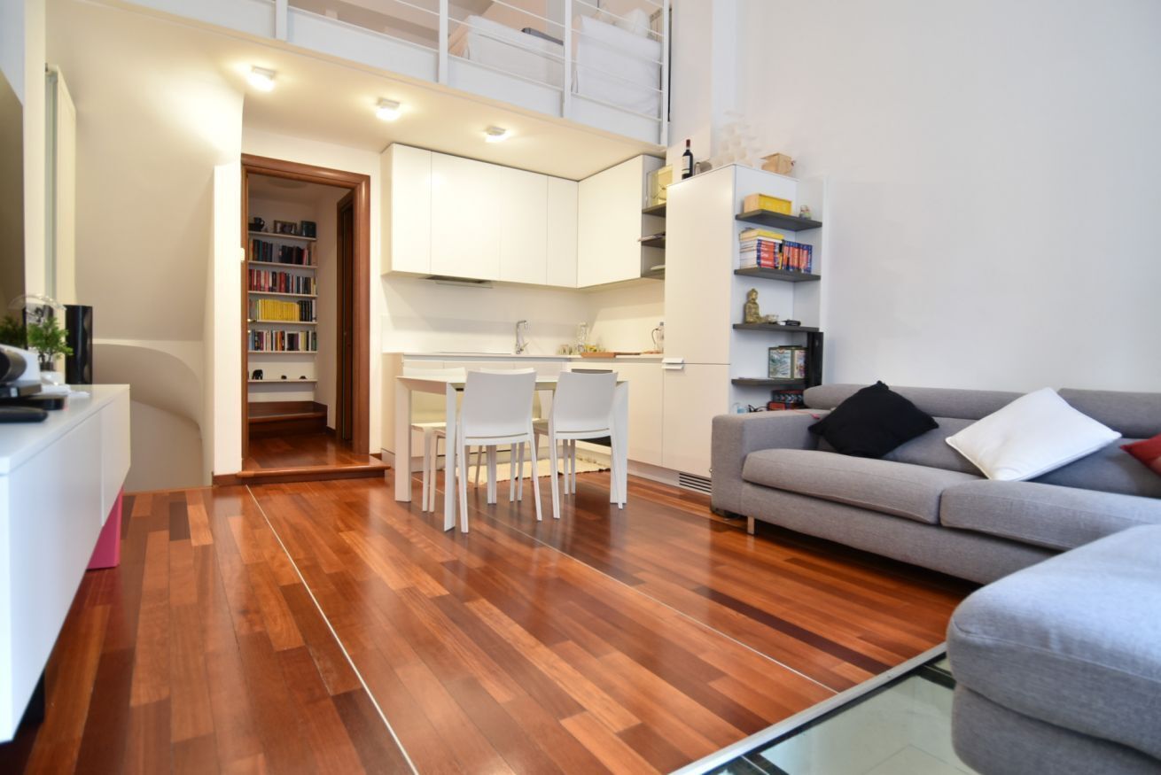 Una sala de estar moderna con un sofá gris, un comedor con sillas blancas y una cocina con gabinetes blancos.