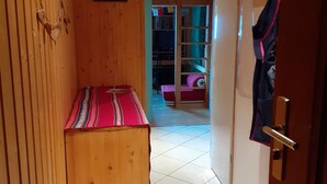 Passadís estret amb parets de fusta, una banca, una porta amb un abric penjant i una visió d'una habitació amb un llit i un armari.