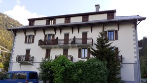 Un edifici amb balcons, una furgoneta blava estacionada i vegetació al voltant.