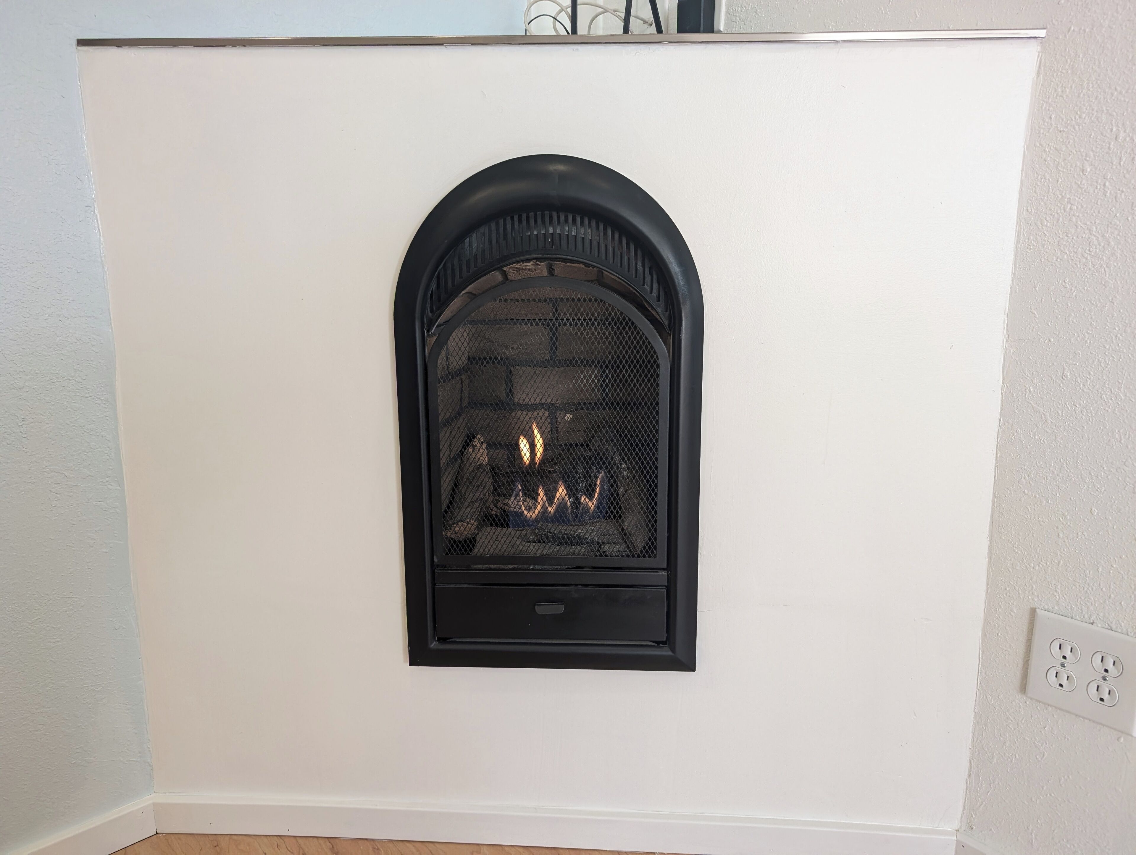 A white wall with a black fireplace, a visible electrical outlet, and a wooden floor.