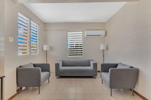 Een kamer met twee grijze banken, een raam met zonwering, een airconditioning en een lamp.