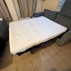 Een compact bed met een wit matras in een kamer met betegelde vloer en een grijze bank.