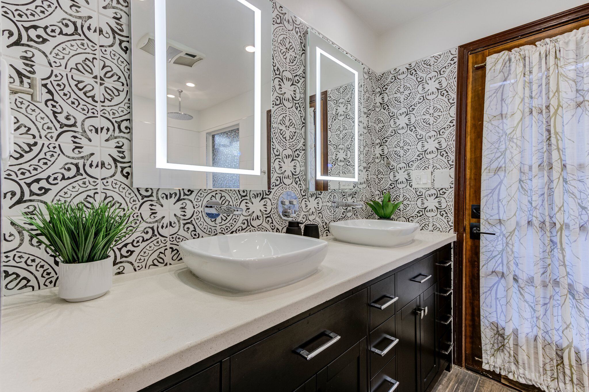 A bathroom with patterned wallpaper, two sinks, a potted plant, and a mirror with integrated lighting.