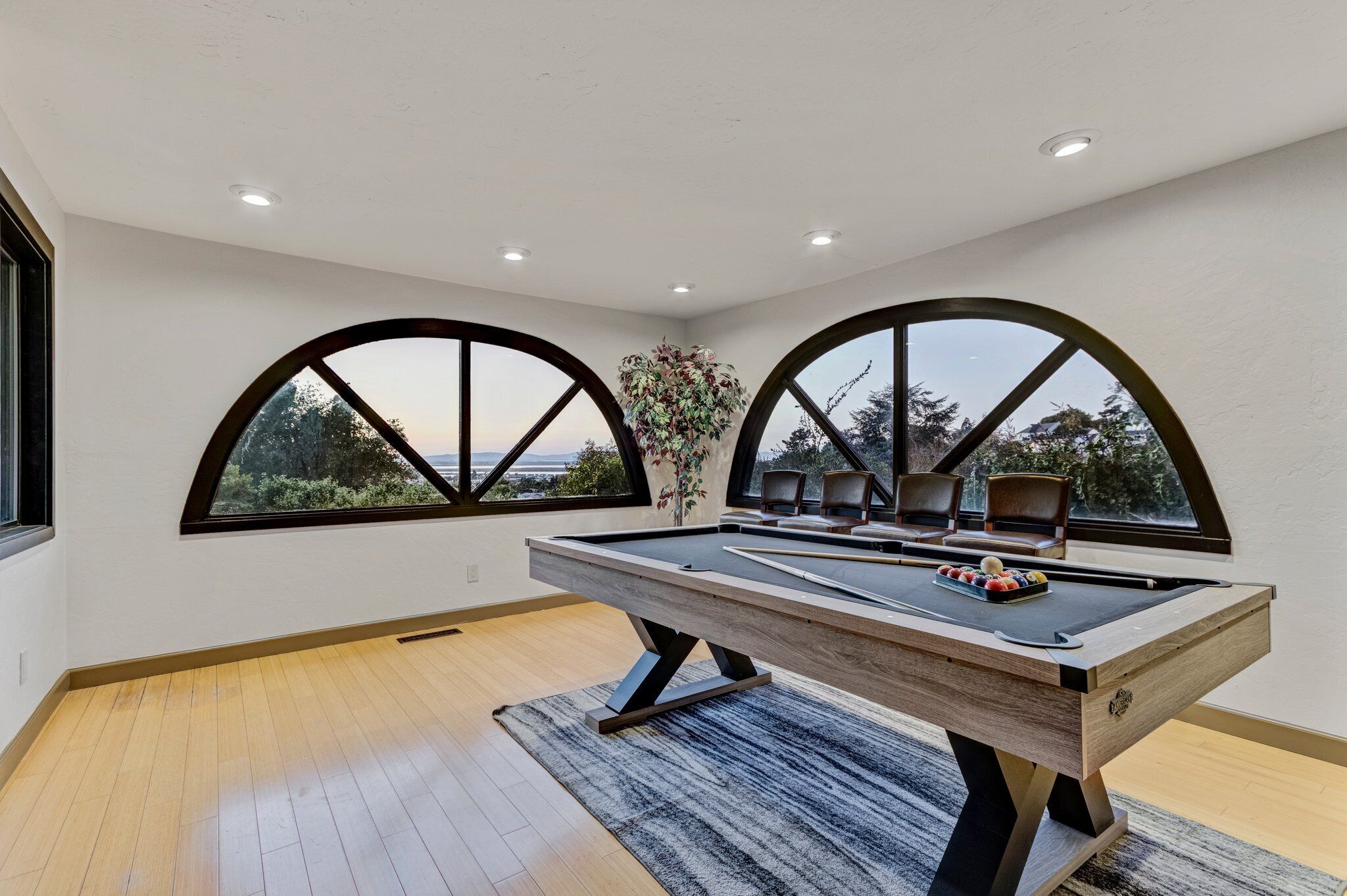 Una mesa de billar en una habitación con dos amplias ventanas arqueadas, una planta en maceta y vistas a árboles y montañas.