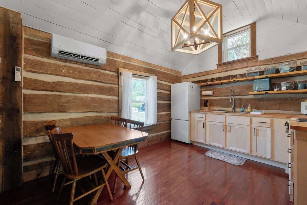 Interior rústico de una cabaña con una mesa de comedor de madera, sillas, un refrigerador y una zona de cocina con estantes y armarios de madera.