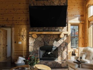 L’intérieur d’une cabane en rondins, avec une cheminée en pierre, un téléviseur fixé au mur et un coin salon confortable.