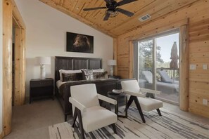 A log cabin bedroom with a bed, two chairs, a ceiling fan, and a large window.