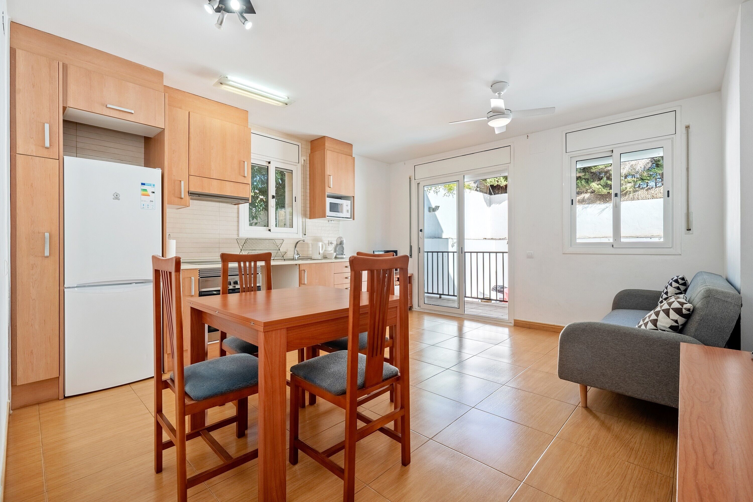 A modern kitchen with wooden cabinets, a dining table with chairs, a grey sofa, and a sliding glass door leading to a balcony.
