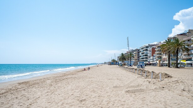 Ein Strand mit sandigem Ufer, blauem Himmel und Gebäuden im Hintergrund.
