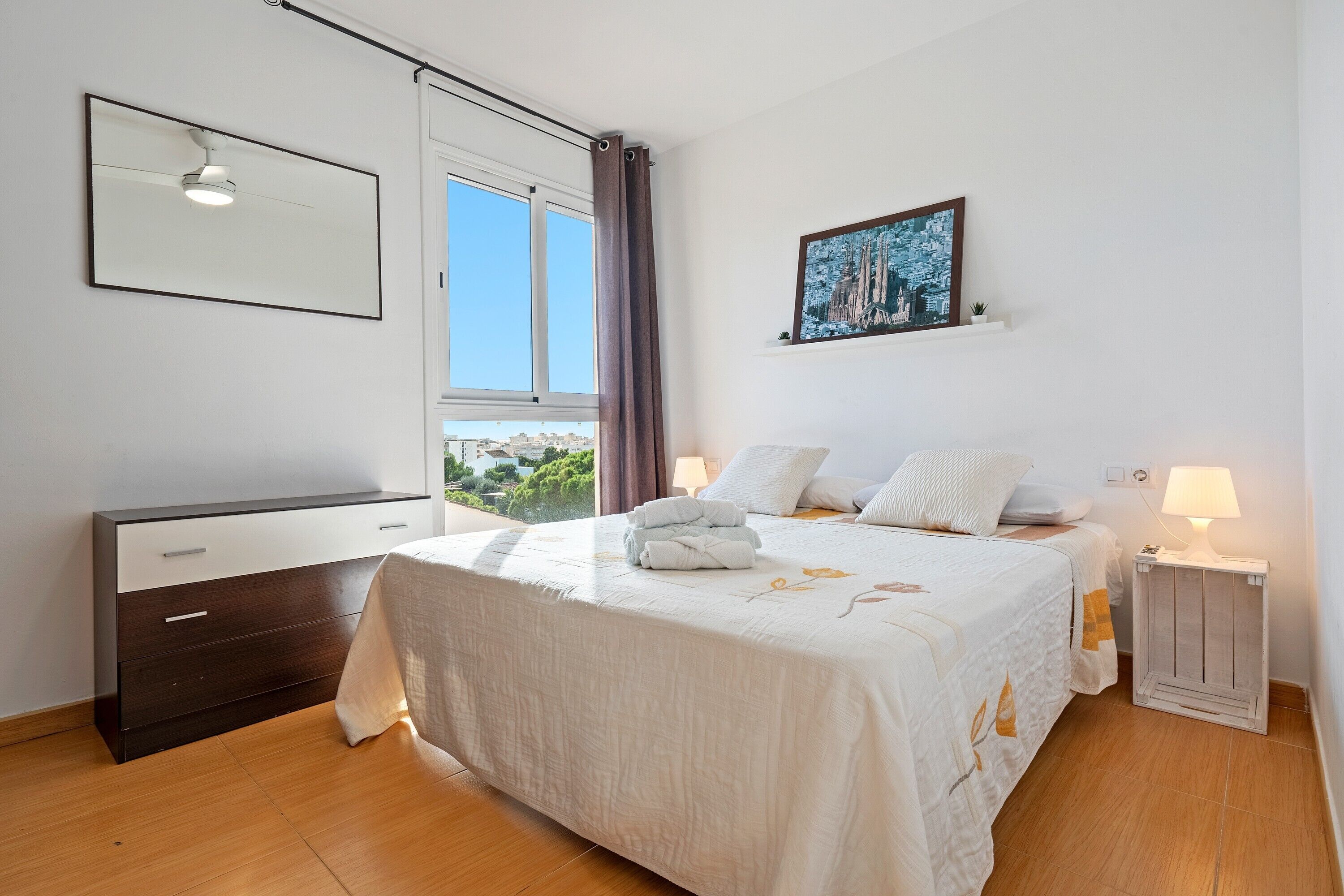 A bedroom with a bed, bedside tables, a dresser, a window with curtains, a painting, and a view of the outdoors.