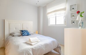 A neatly made bed with a white duvet, a blue patterned pillow, and a folded white towel. There is a bedside table with a vase containing a single red rose.