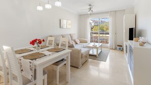 A modern living room with a dining area, a sofa set, a flat-screen TV, and a balcony with a view. - Benalmádena-Arenal Golf Apartment with Shared Pool, Wi-Fi, and Air Conditioning (Benalmádena)