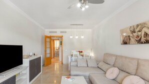 A modern living room with a flat-screen TV, a sofa, a coffee table, and a dining area in the background. - Benalmádena-Arenal Golf Apartment with Shared Pool, Wi-Fi, and Air Conditioning (Benalmádena)