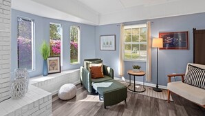 Un salon moderne avec un canapé, un fauteuil et un pouf, de grandes fenêtres donnant sur un paysage verdoyant et un lampadaire.