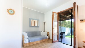 Una habitación con piso de madera, un sofá gris, un reloj de pared, un espejo enmarcado y un vidrio corredizo que da a un área exterior.
