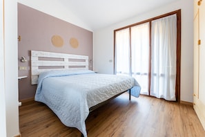 Une chambre avec un plancher en bois, un lit blanc avec une tête de lit et une grande fenêtre avec des rideaux.