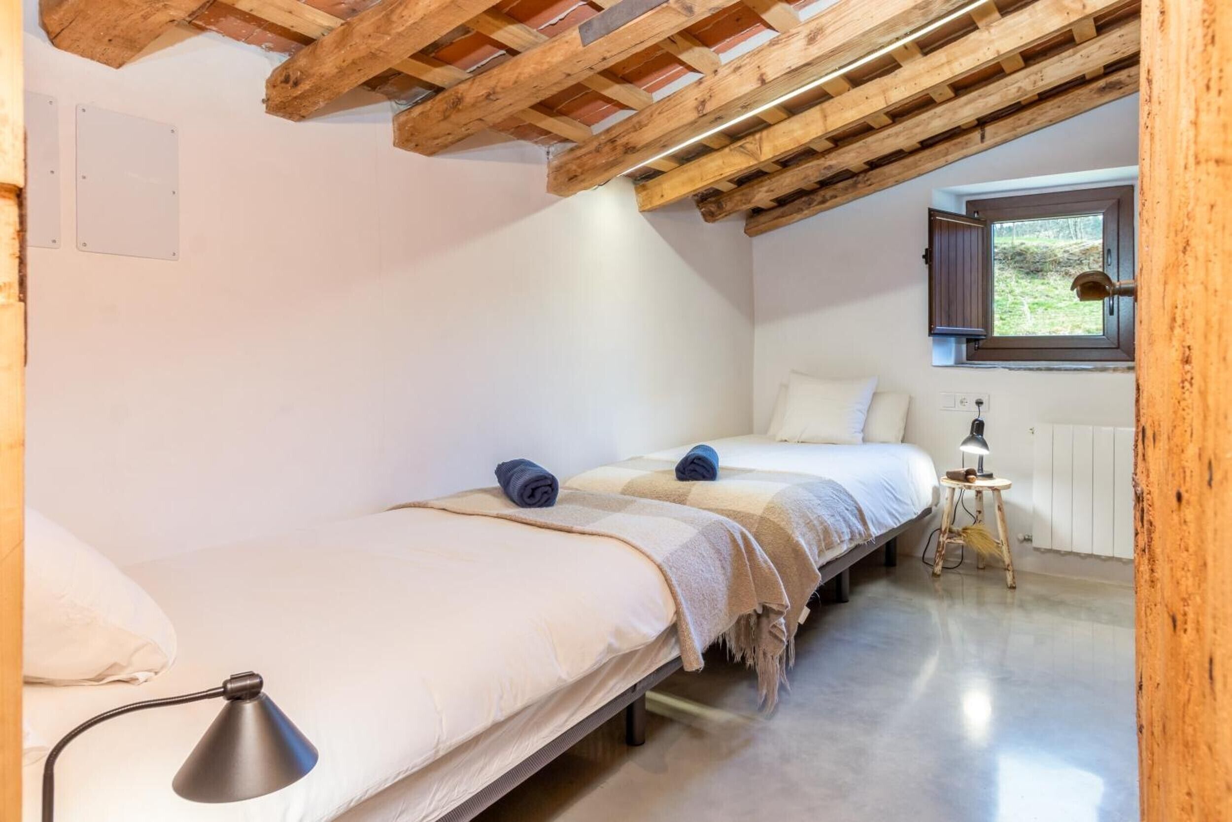 A bedroom with two beds, a window with a view, a lamp on a bedside table, and a wooden ceiling structure.