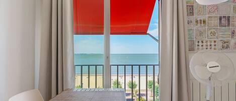 A balcony with a view of the sea, a table, a chair, a fan, and a wall with various pictures.