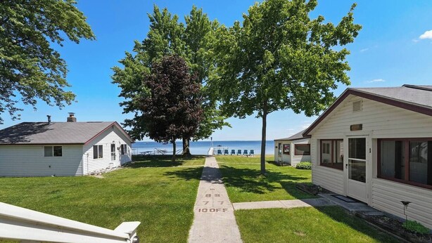 Une rangée de cabanes bordée d’un sentier herbeux menant à une plage, un grand arbre au centre et un ciel bleu dégagé.