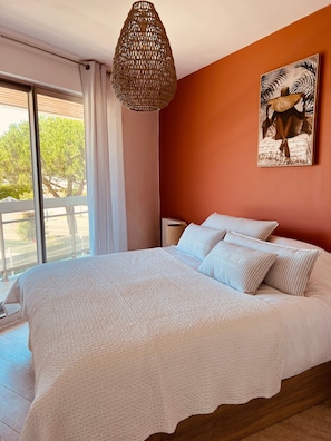 A bedroom with a large bed, a hanging light fixture, a painting on the wall, and a view of the outdoors through a window.