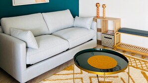 A modern living room with a teal accent wall, a light grey sofa, a coffee table with a glass top, and a patterned rug.