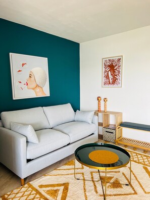 A modern living room with a teal accent wall, a light grey sofa, a coffee table with a glass top, and a patterned rug.