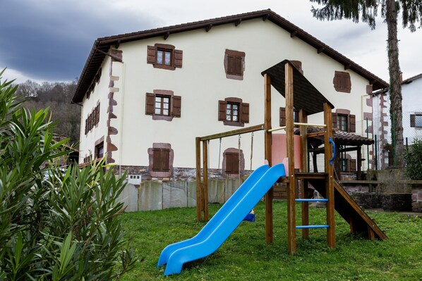 Ein Spielplatz mit einer blauen Rutsche und einer hölzernen Kletterkonstruktion vor einem Gebäude mit vielen Fenstern und einem geneigten Dach.