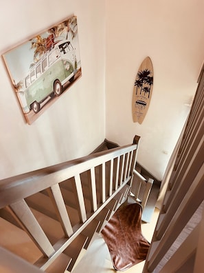 Un escalier avec une rampe en bois, une planche de surf accrochée au mur et un tableau représentant une voiture ancienne.