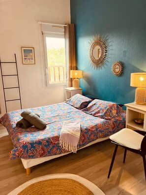 Un dormitorio con cama, dos lámparas de noche, una silla, una mesita, un reloj de pared, un cuadro enmarco y una ventana con cortinas.