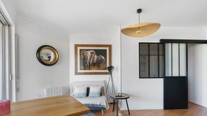 Una sala moderna con una mesa de comedor de madera, un sofá, una lámpara de pie, un cuadro enmarcado de un elefante y un amplio ventanal con persianas.