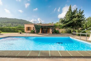 Une piscine avec un espace patio, un petit bâtiment doté d’un toit en tuiles et une colline en arrière-plan.